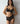 A woman modeling a black lace lingerie set against a plain light gray wall. The set includes a sheer mesh and lace underwire bra with gold-tone hardware and matching high-waisted lace panties.