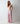 A woman with red hair stands barefoot in a studio setting, wearing a light pink bralette and matching pink vertically striped pajama pants against a plain light gray background.