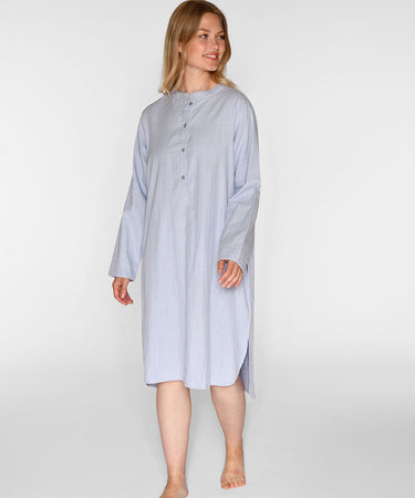 A woman modeling a light blue and white vertically striped long-sleeve nightshirt or tunic with a buttoned neckline, standing against a plain light gray studio background.